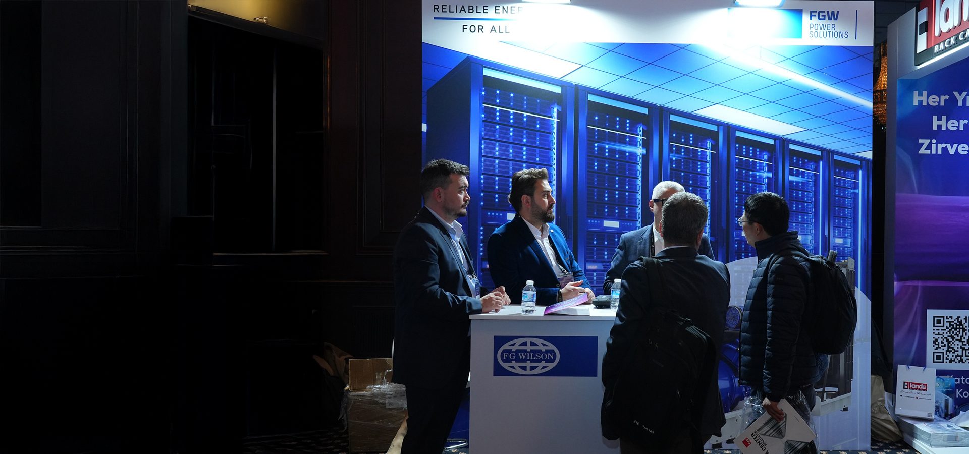 People networking at an FG Wilson tech expo booth with a server rack graphic backdrop.