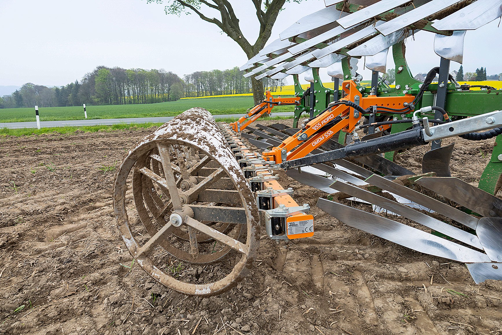 Muddy agricultural roller and plow in a field, beside a road with trees and yellow fields.