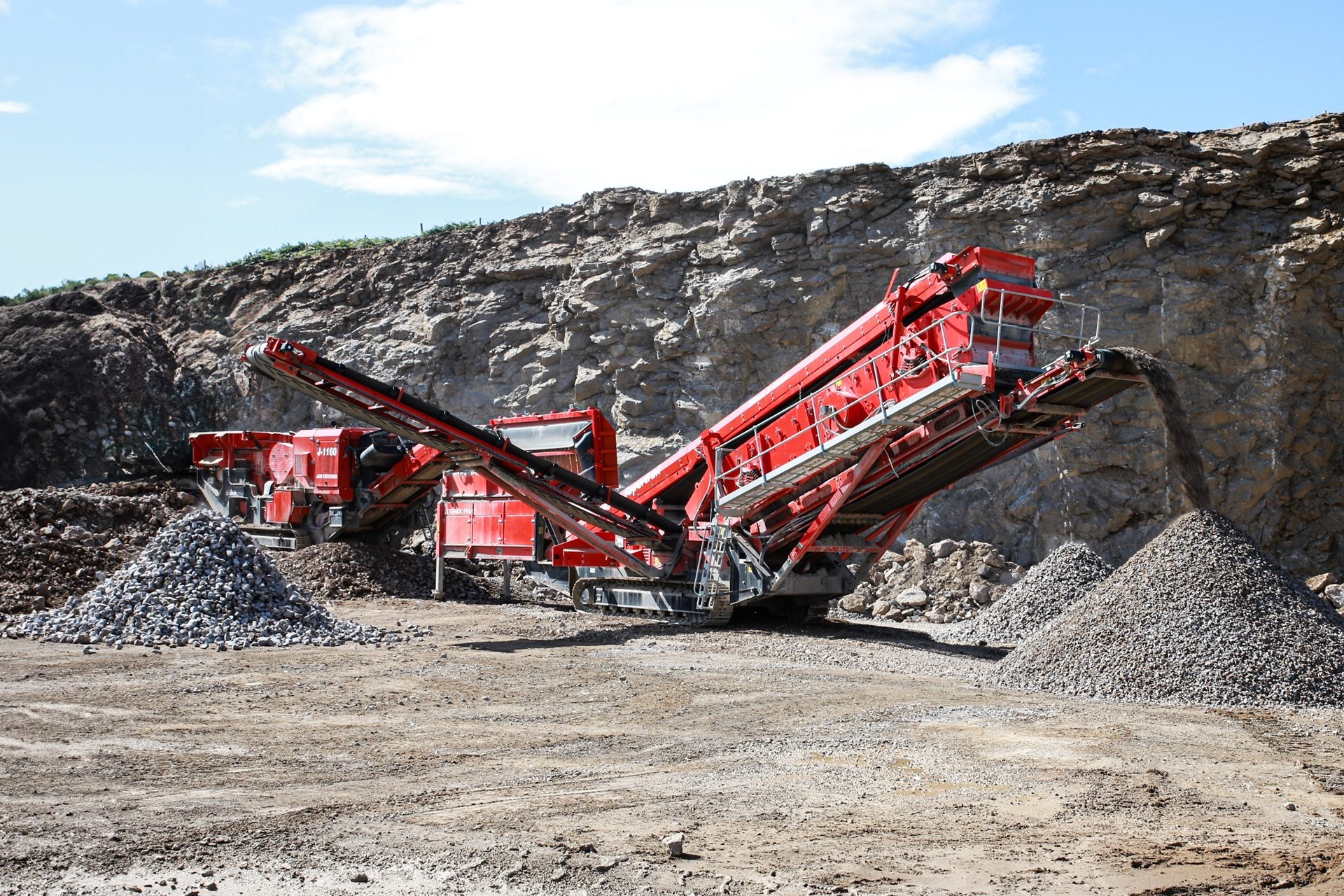 Large red rock crushing machine processing rocks into piles of gravel in a quarry under a blue sky.