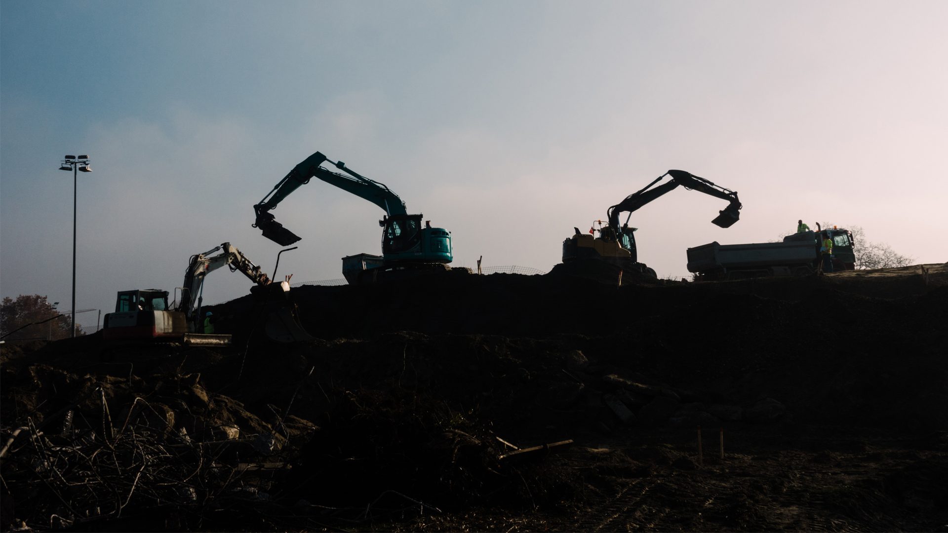 Silhouetted construction site with excavators, a dump truck, and workers against a bright sky.