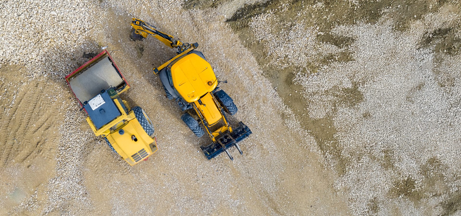 Aerial view of a yellow road roller and a yellow backhoe on a gravel construction site.