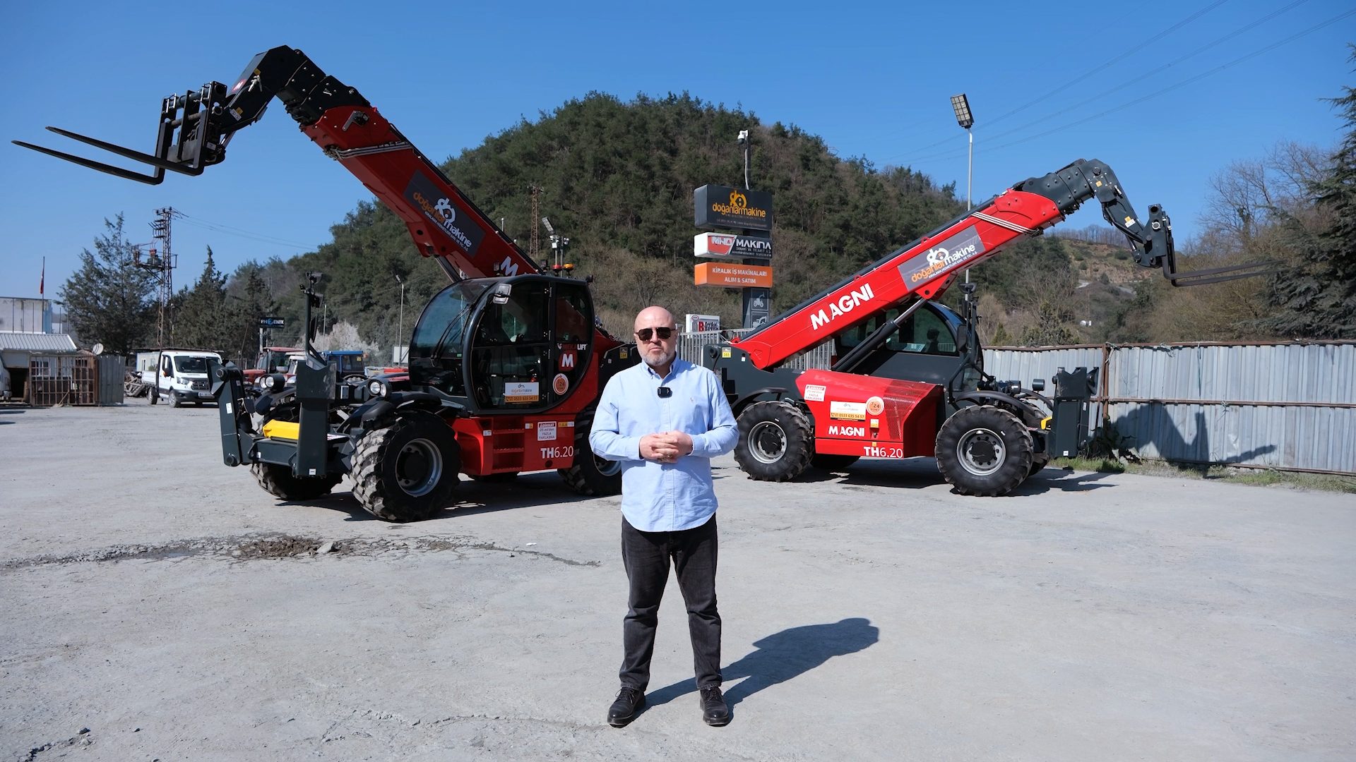 A man stands between two red Magni telehandlers in an outdoor industrial setting.