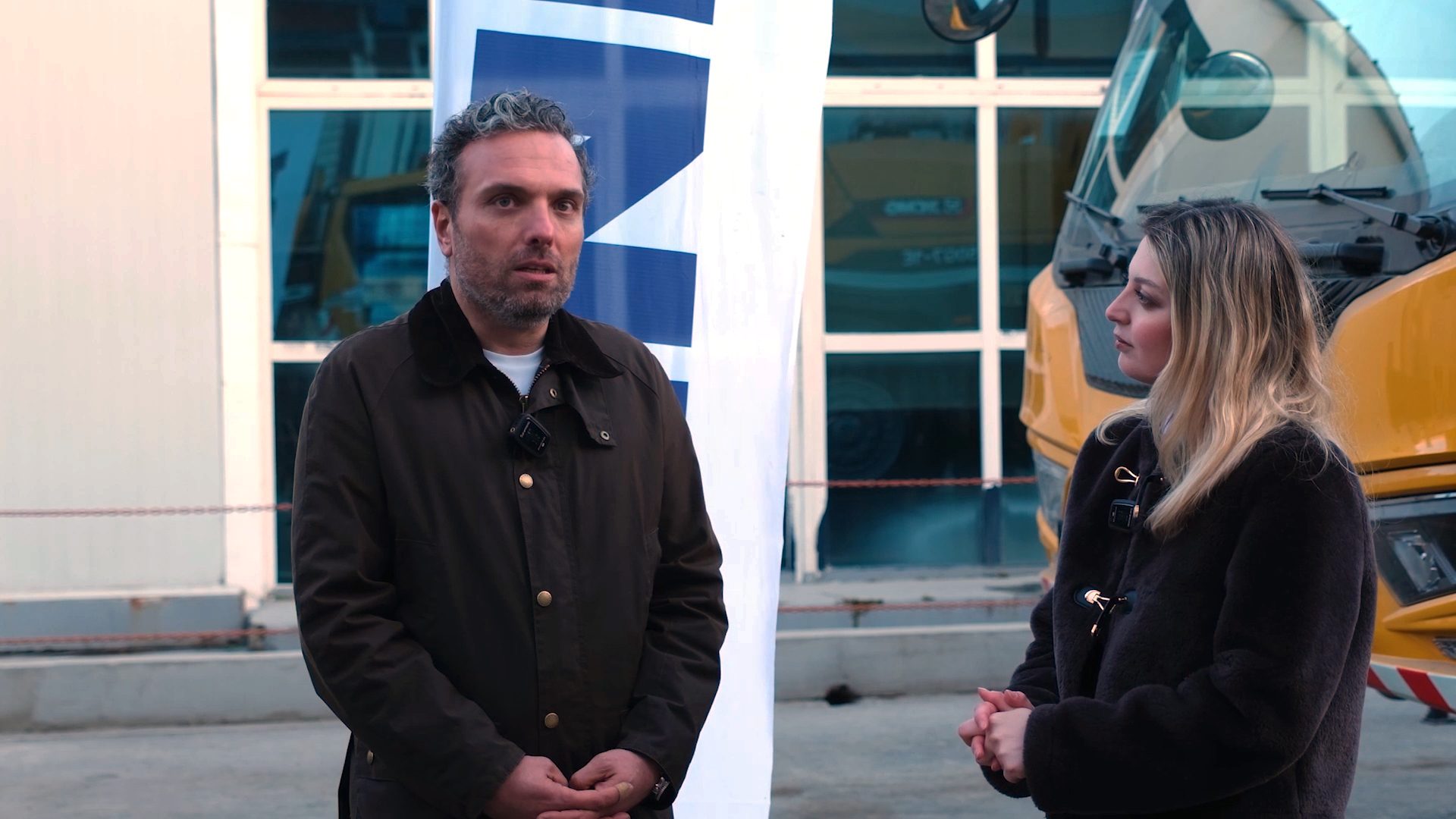A man and a woman in outdoor jackets are standing, appearing to have a conversation.