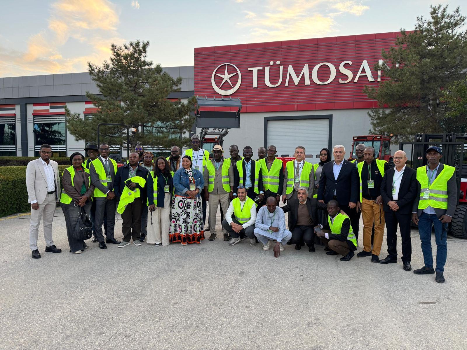A diverse group of people, many in high-visibility vests, pose in front of the TÜMOSAN building.