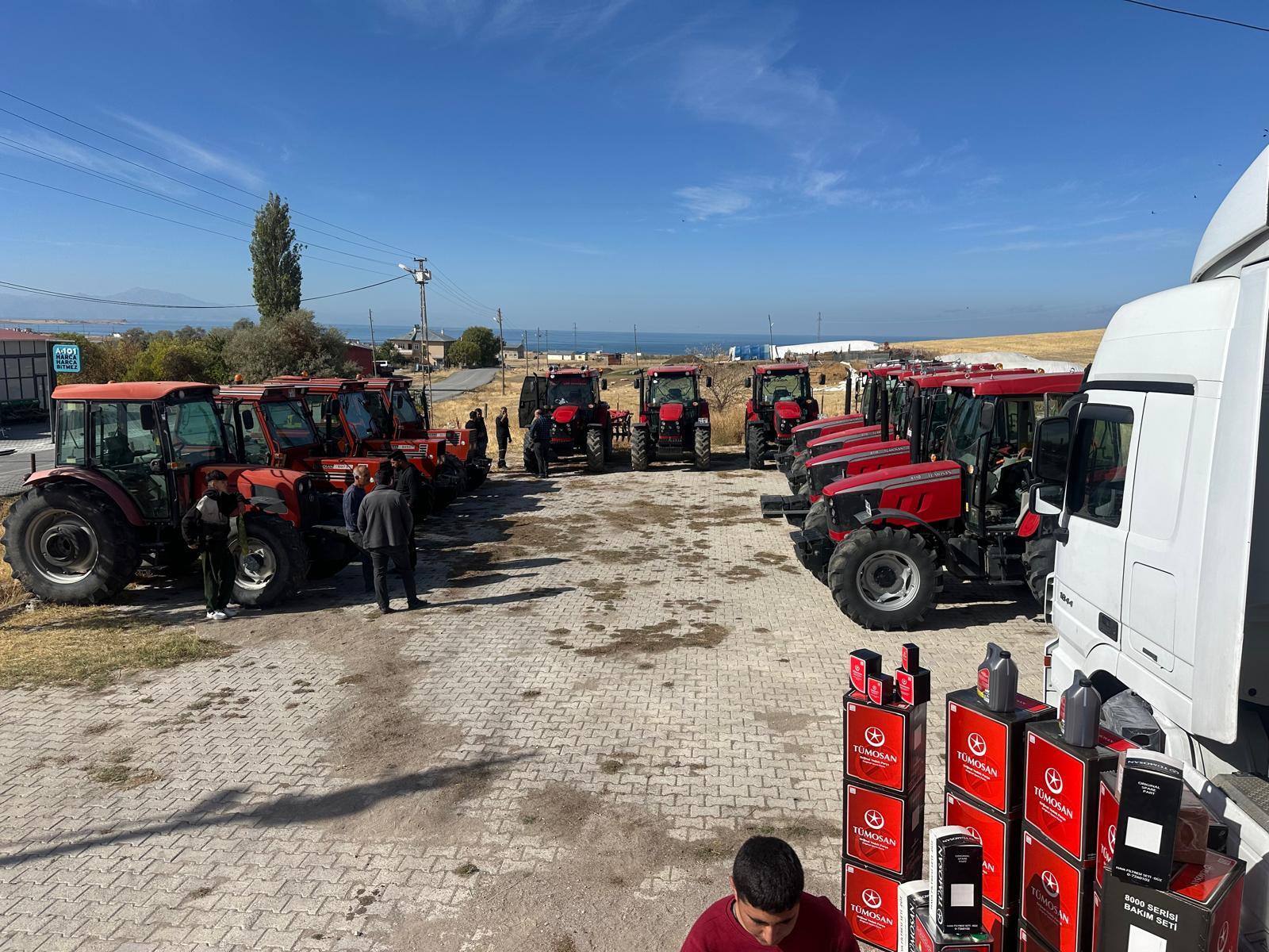 Many red tractors, people, a truck with Tümosan parts, and distant water under a blue sky.