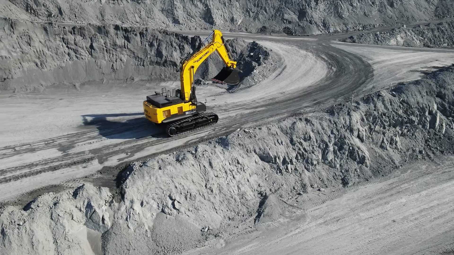 Yellow excavator on a curved road in a gray open-pit mine.
