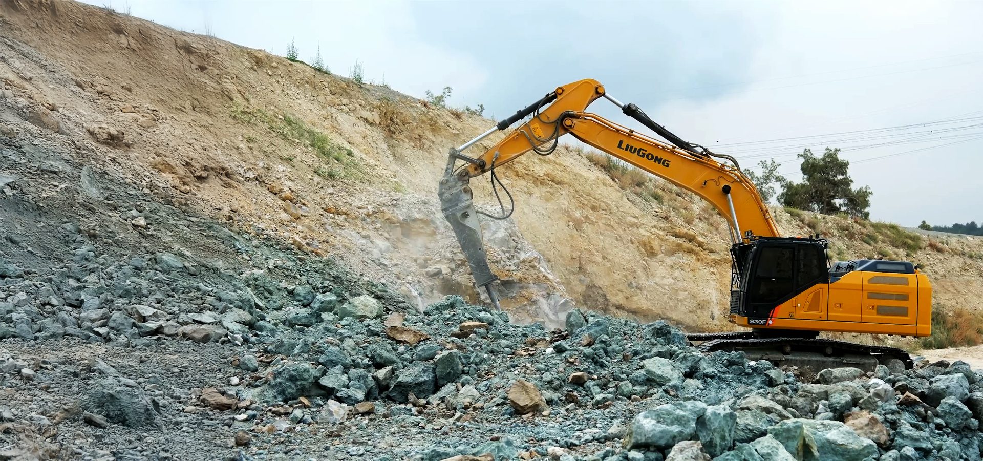 Yellow excavator with hydraulic hammer breaking blue-green rocks on a hillside.