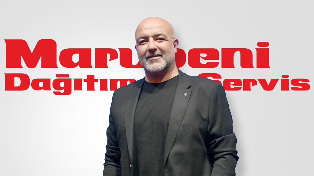 A bald man with a beard, wearing a black suit jacket and t-shirt, smiles at the camera in front of a red logo.