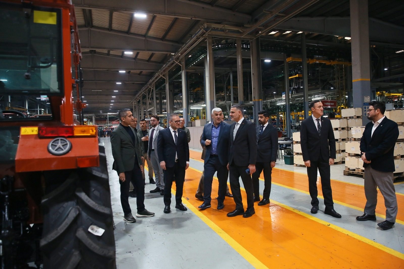 Men tour a factory, walking on orange-marked paths, with a red tractor in the foreground.