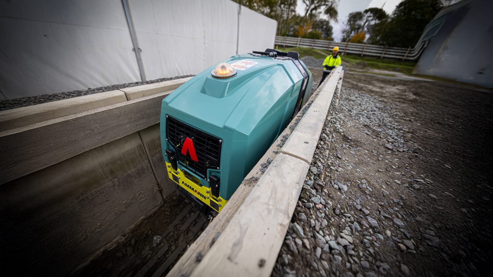 Teal trench compactor machine in a narrow trench with a worker in the background.