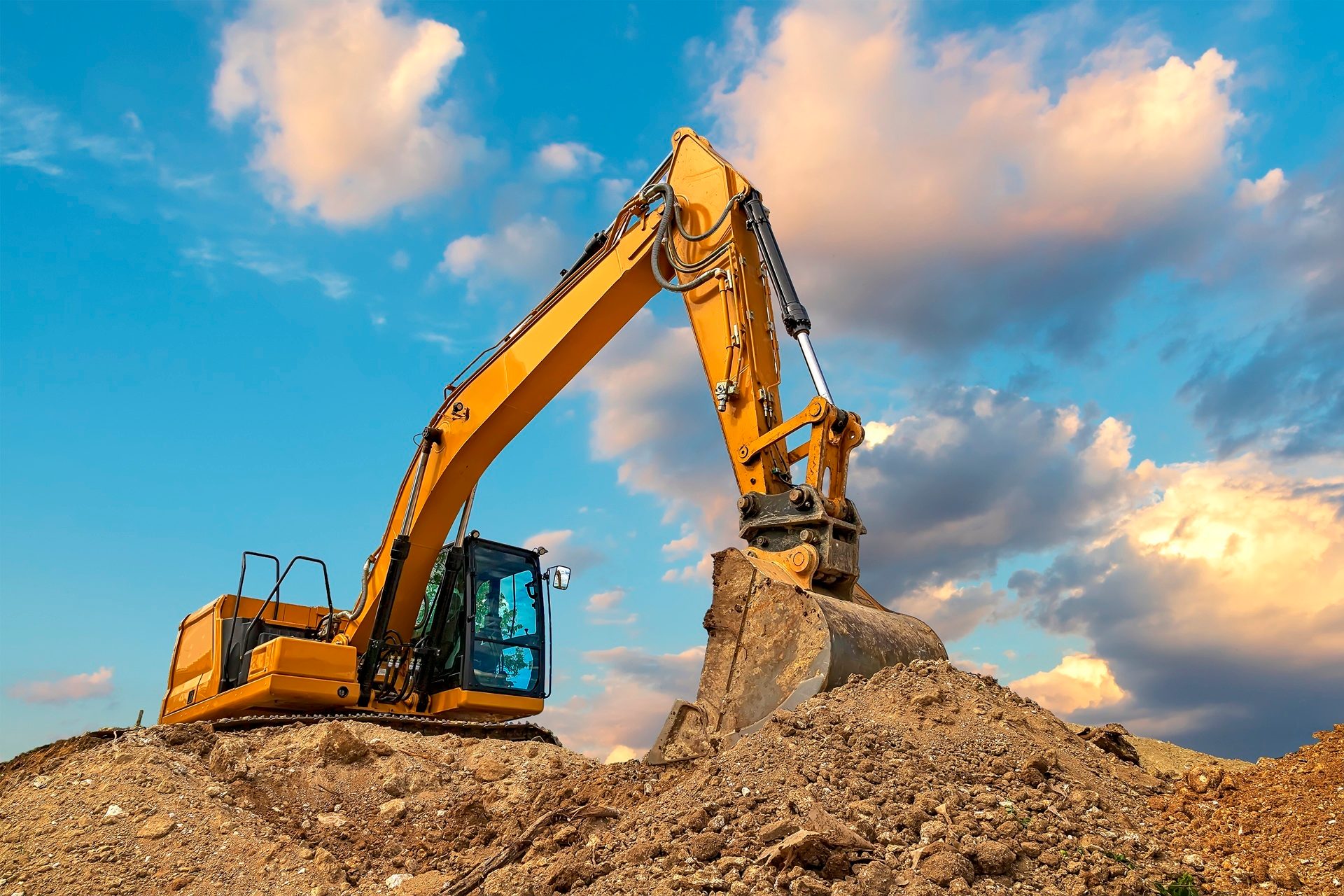 Yellow excavator on a dirt mound under a cloudy blue sky.