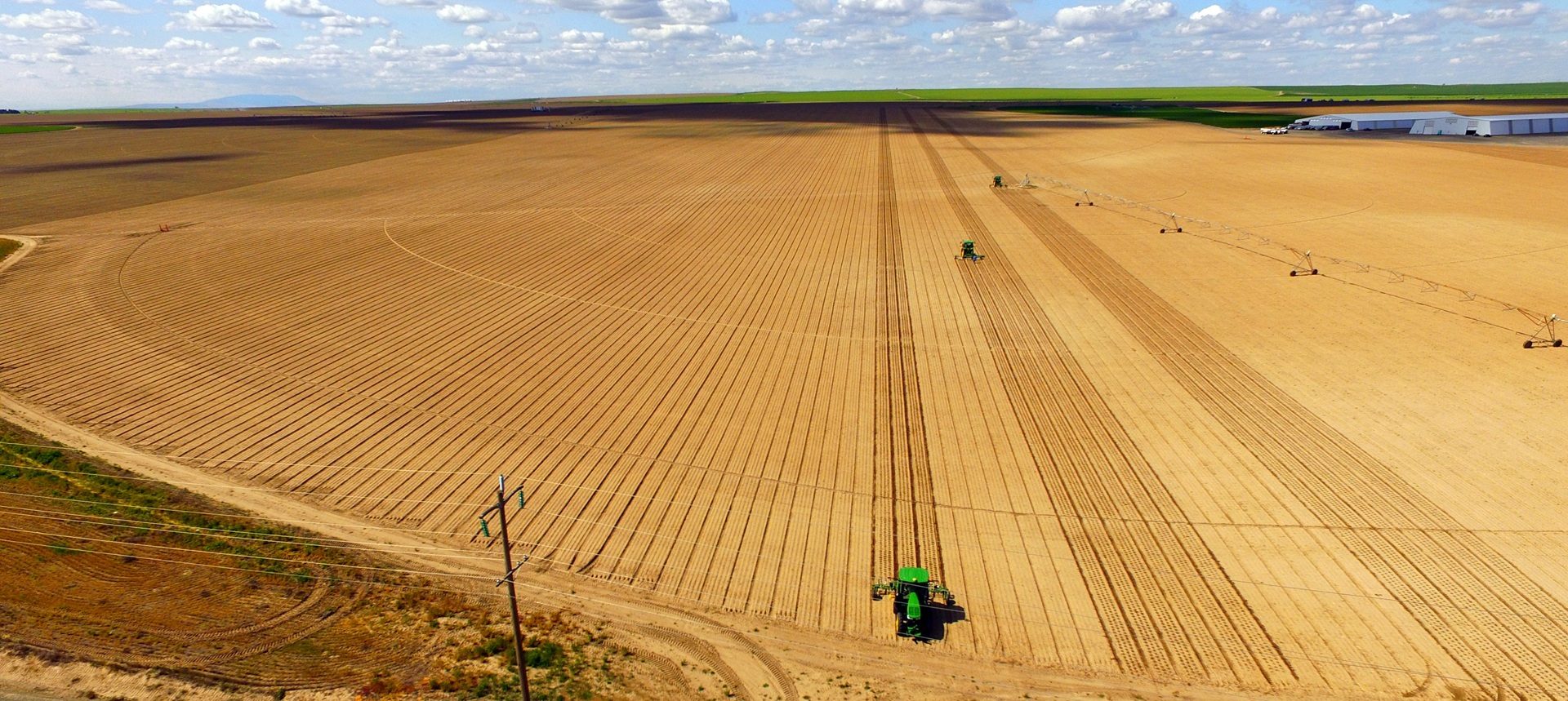 Aerial view of a vast tilled field with tractors working, irrigation, distant buildings, and a cloudy sky.
