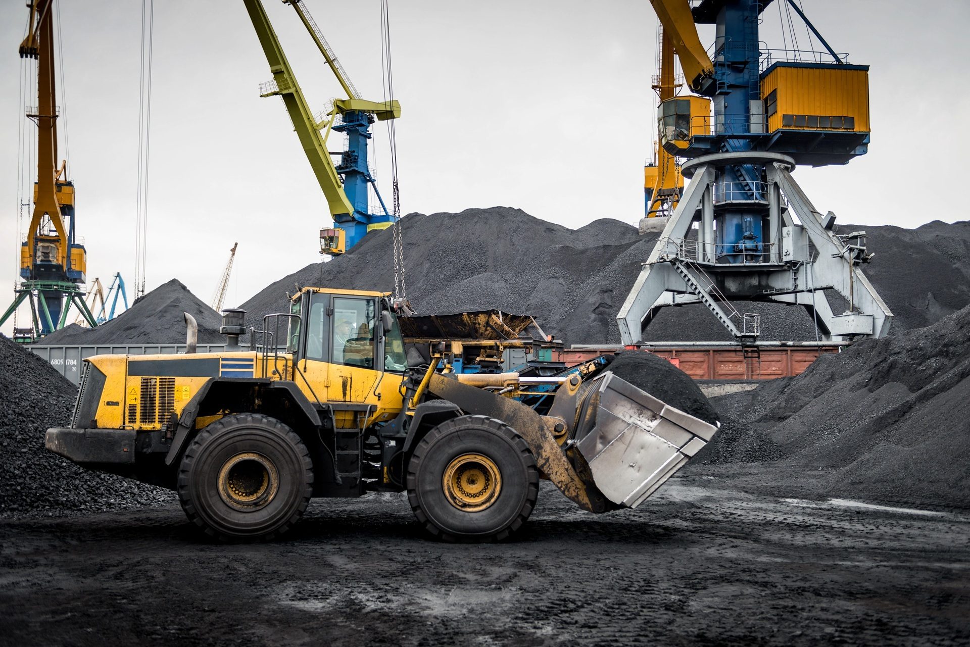 Yellow front-end loader moving coal amidst large piles and industrial port cranes under a grey sky.