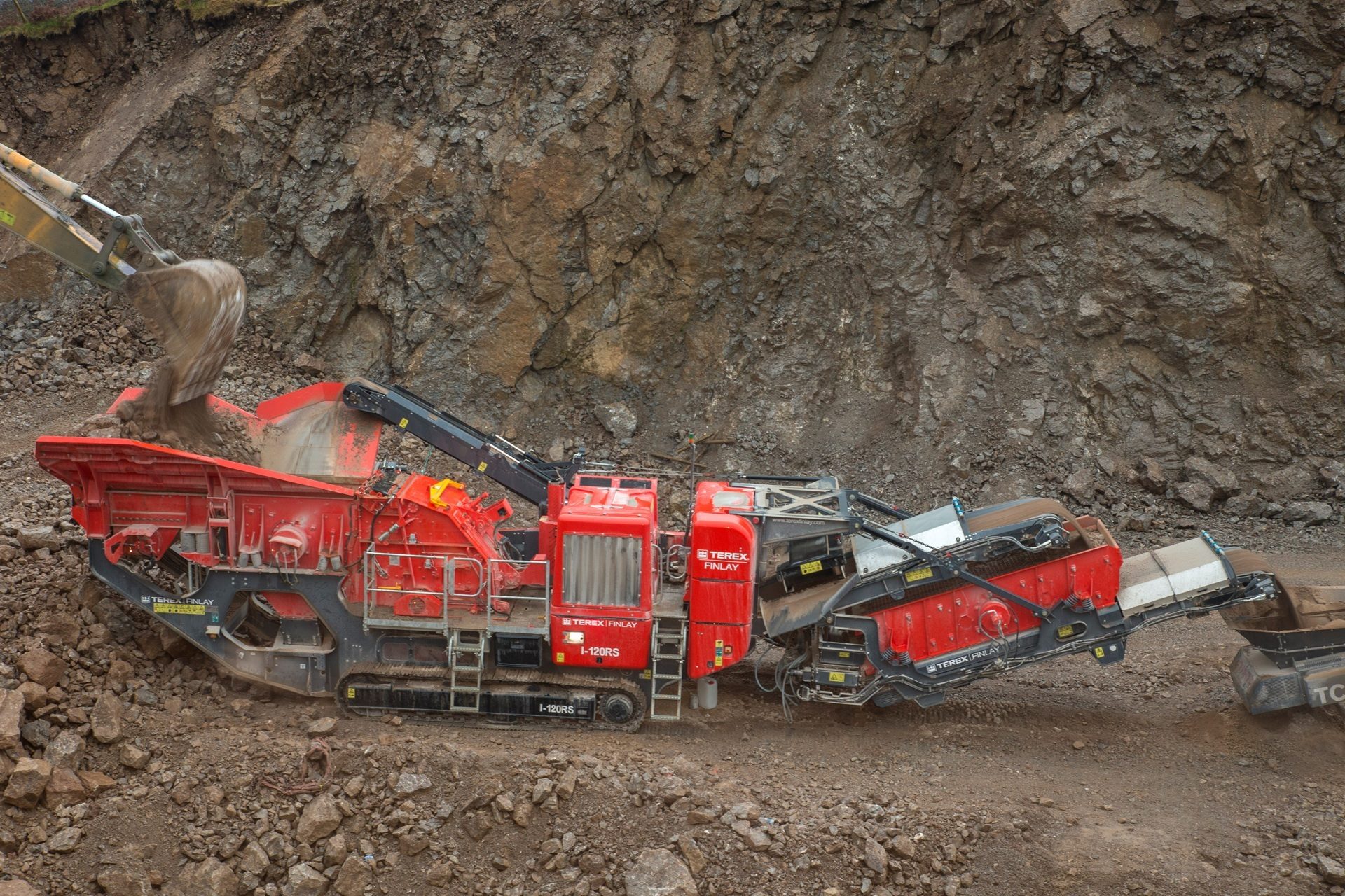 An excavator feeds rocks into a large red Terex Finlay I-120RS mobile crusher in a quarry.