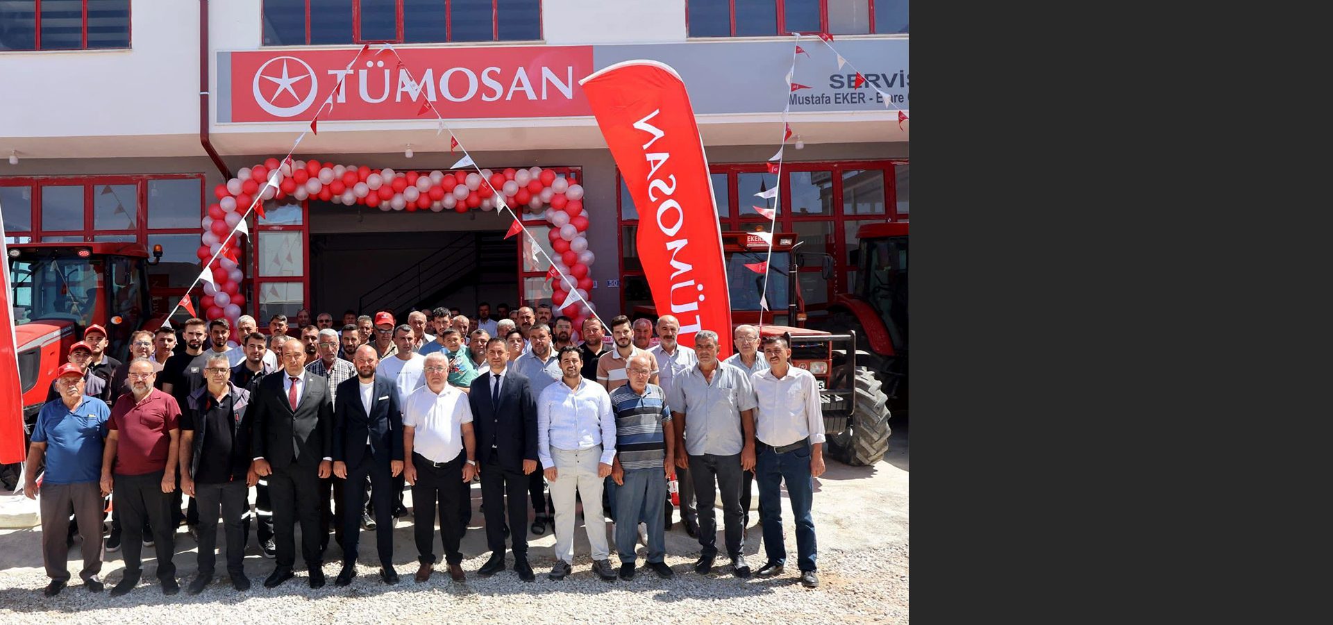 A group of men stand in front of a TÜMOSAN dealership, decorated with balloons and flags, with tractors.