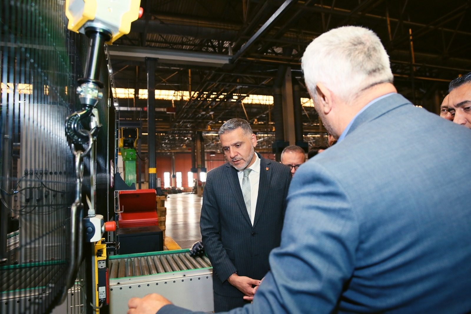 Man in a suit inspects factory machinery, accompanied by other men.