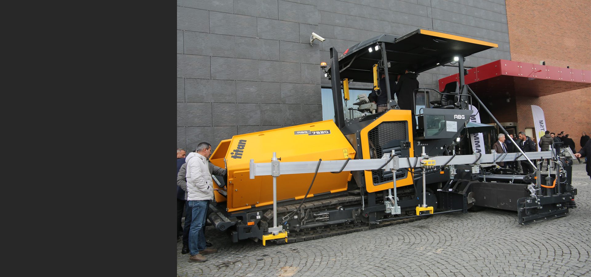 A large yellow Titan ABG 7820 road paver machine is displayed outdoors with people observing it.