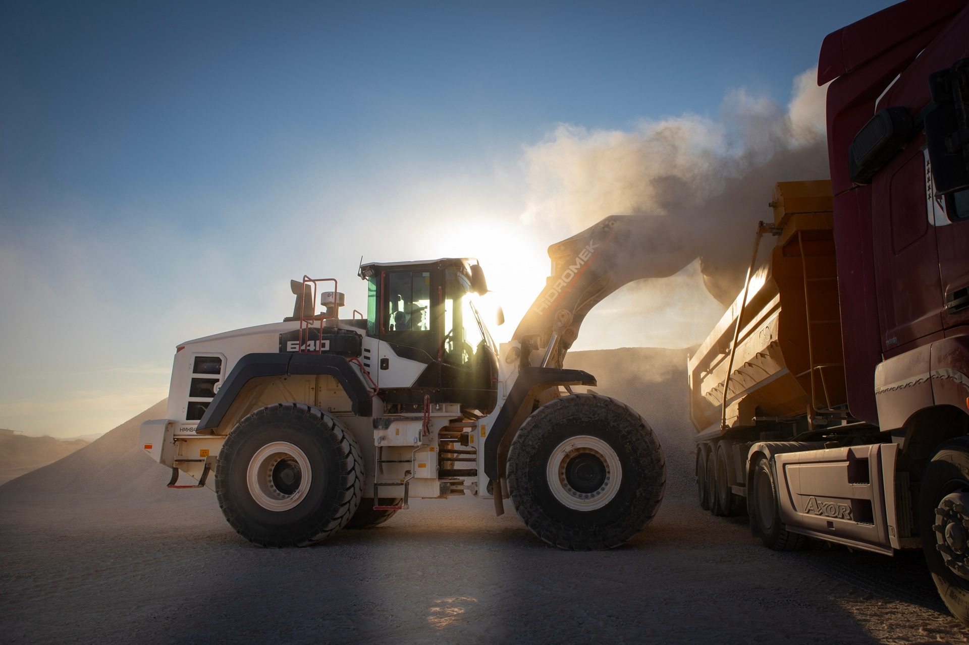 Front-end loader fills dump truck under bright sun, dust billowing.