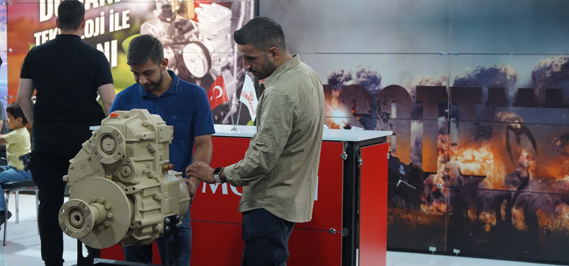 Two men examine an engine at an event, with a backdrop showing explosions and smoke.
