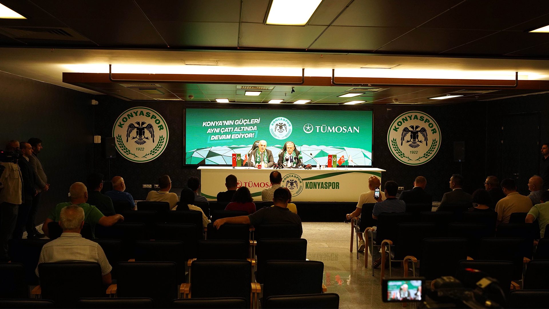 Two men speaking at a Konyaspor press conference with a large screen, logos, and an audience.