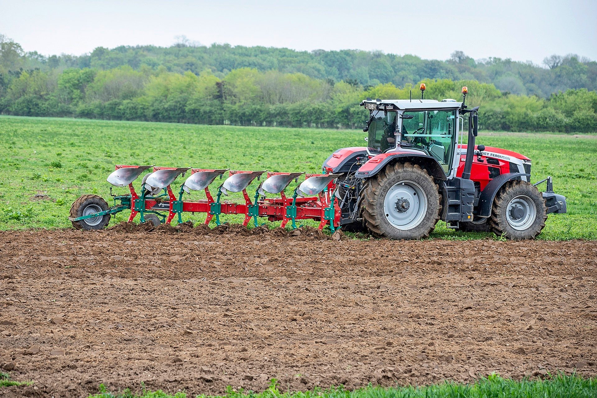 A red and gray tractor with a large plow attachment tilling brown soil in a green field.