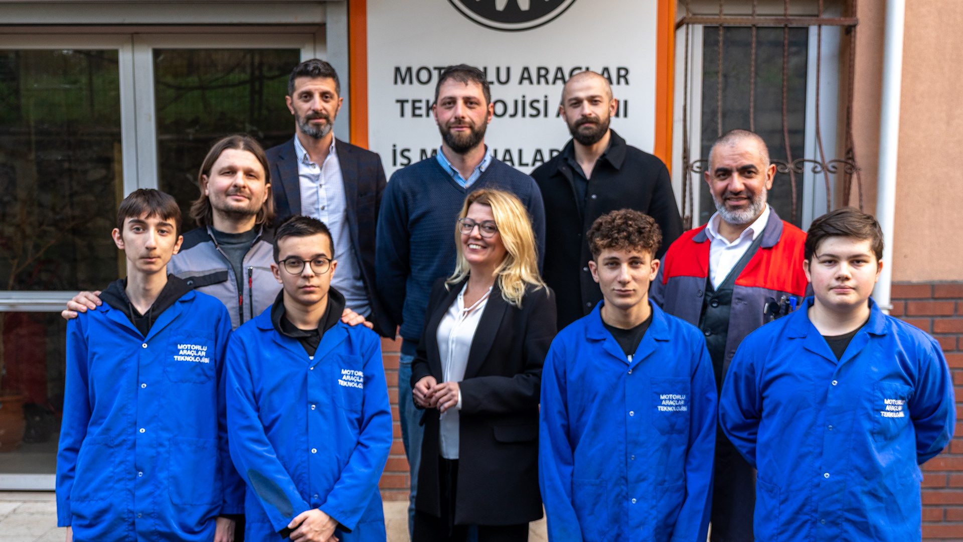 A group of adults and students in blue work coats pose for a photo in front of a building sign.