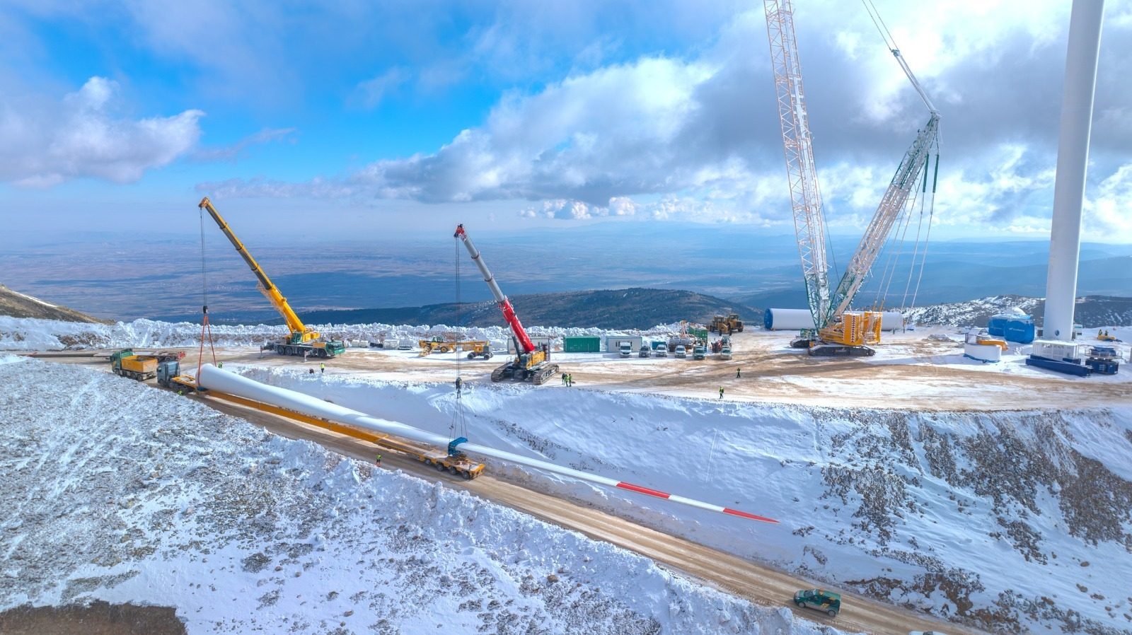 Large wind turbine blade being installed by cranes in a snowy mountain landscape.
