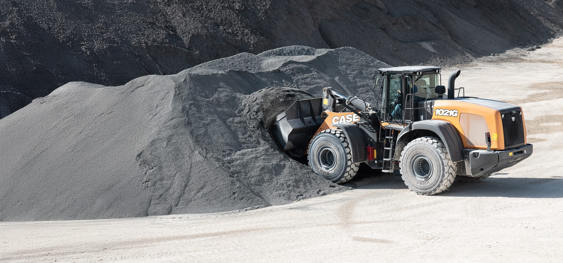 Orange Case 1021G wheel loader digging into a large pile of gray aggregate.