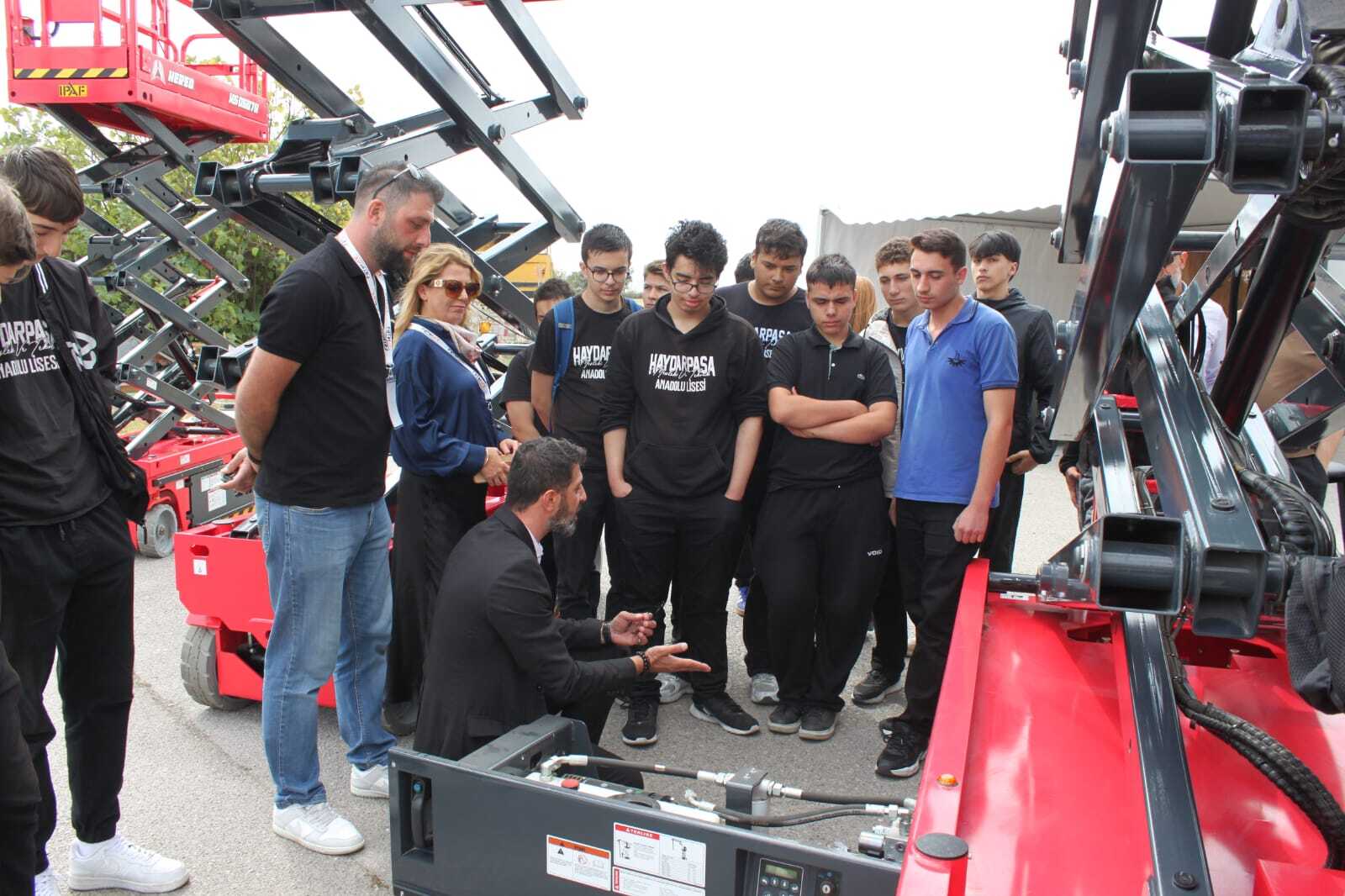 Students and adults gather around a red scissor lift, with an instructor explaining its mechanics.