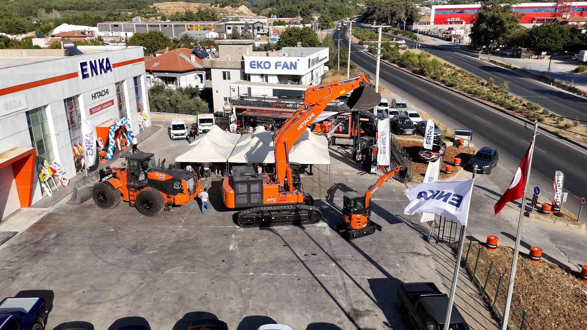 Outdoor showcase of Hitachi construction machinery at an ENKA dealership, with tents, people, and road.