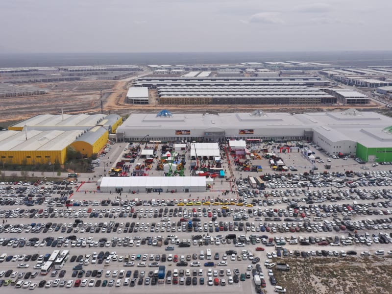 Aerial view of a busy outdoor exhibition with displays, crowds, and a vast parking lot by industrial buildings.
