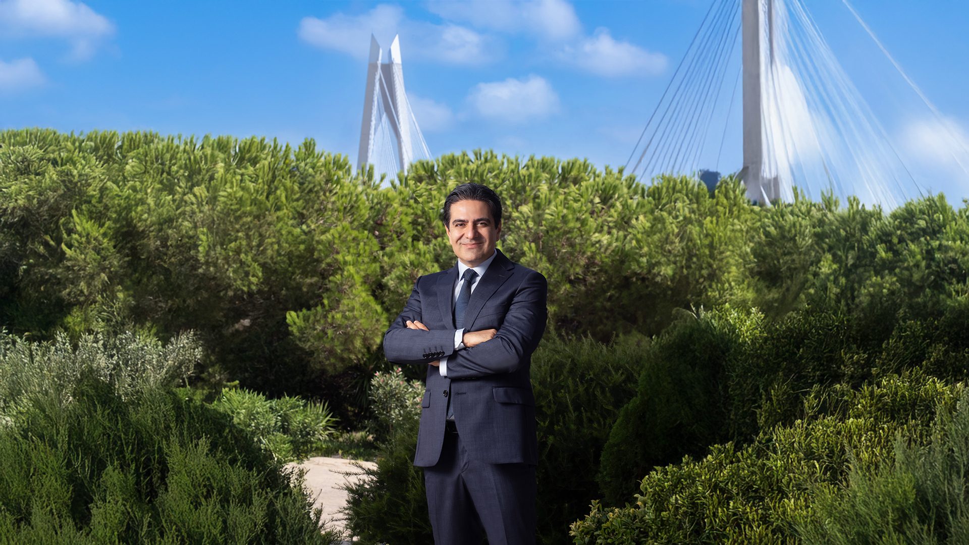 Smiling man in a suit, arms crossed, stands among green trees with a modern white bridge and blue sky.