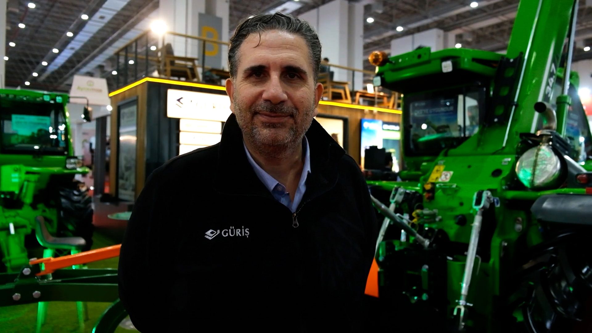 A man in a black jacket smiles at the camera, standing in an exhibition hall with large green machinery.
