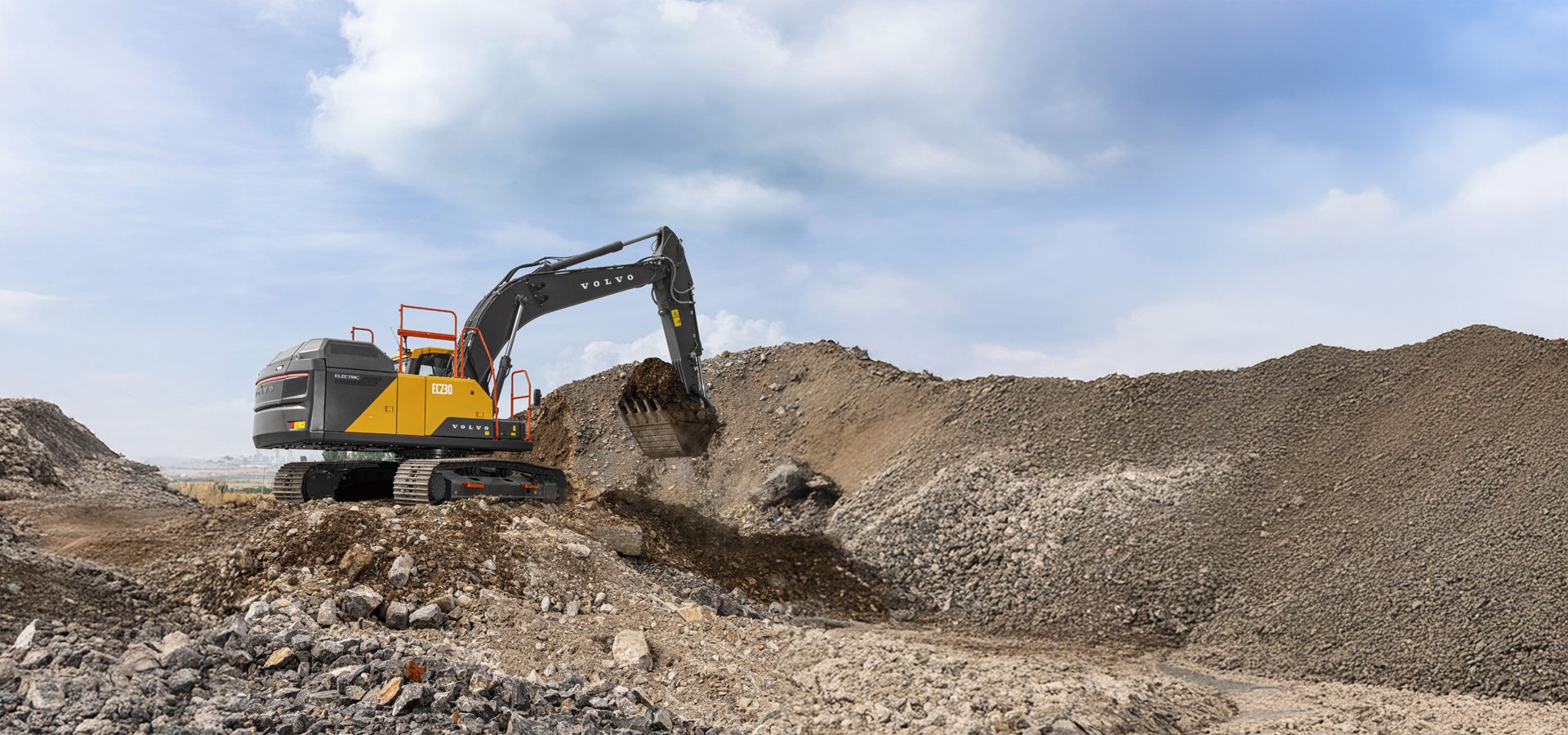 A yellow and black Volvo electric excavator digs into a large pile of gravel and dirt under a cloudy sky.