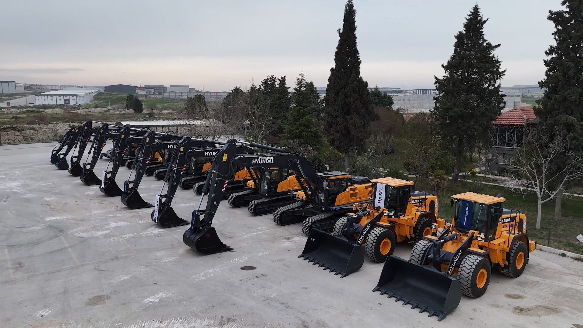 A lineup of new Hyundai excavators and wheel loaders parked on an asphalt lot with industrial buildings in the background.