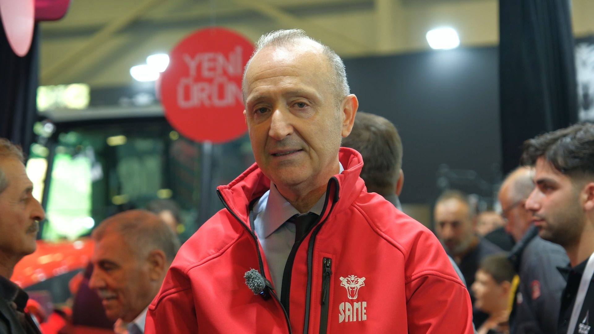 Man in a red jacket with a microphone and "SAME" logo, speaking at an event with others.