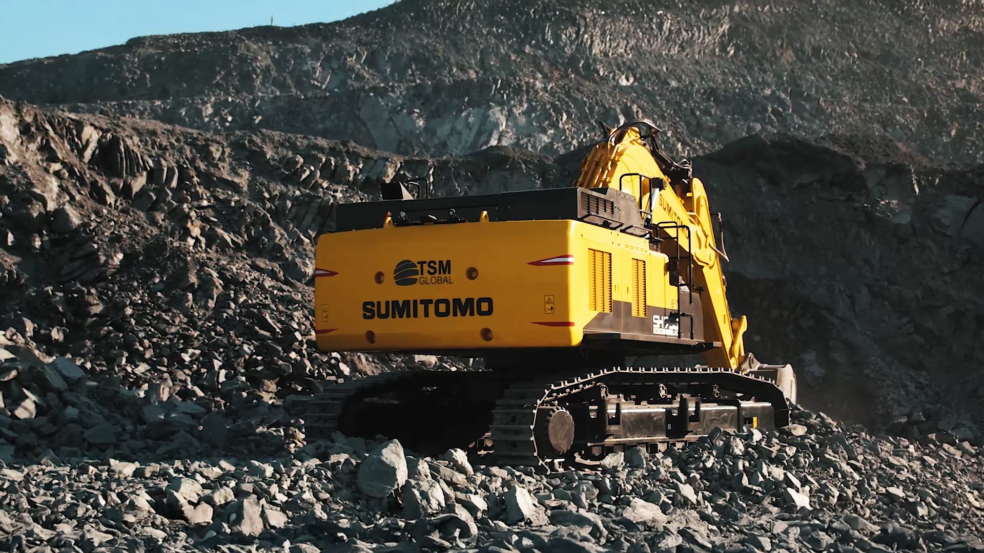 Yellow Sumitomo excavator on a rocky mountainside, seen from the rear.