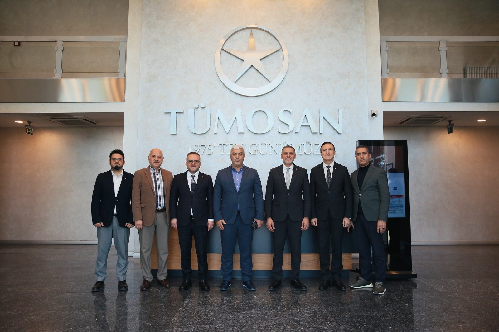 Seven men stand in front of a wall with the "TÜMOSAN" logo and name.