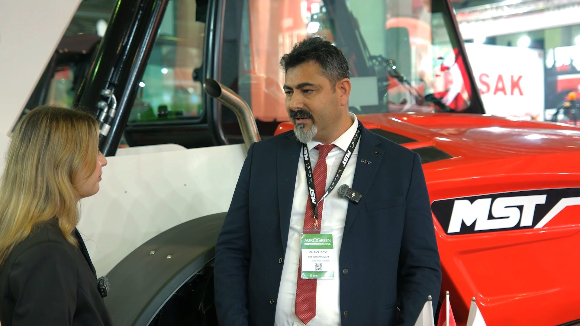 A man in a suit is interviewed by a woman next to a red MST tractor at an Agrogreen event.