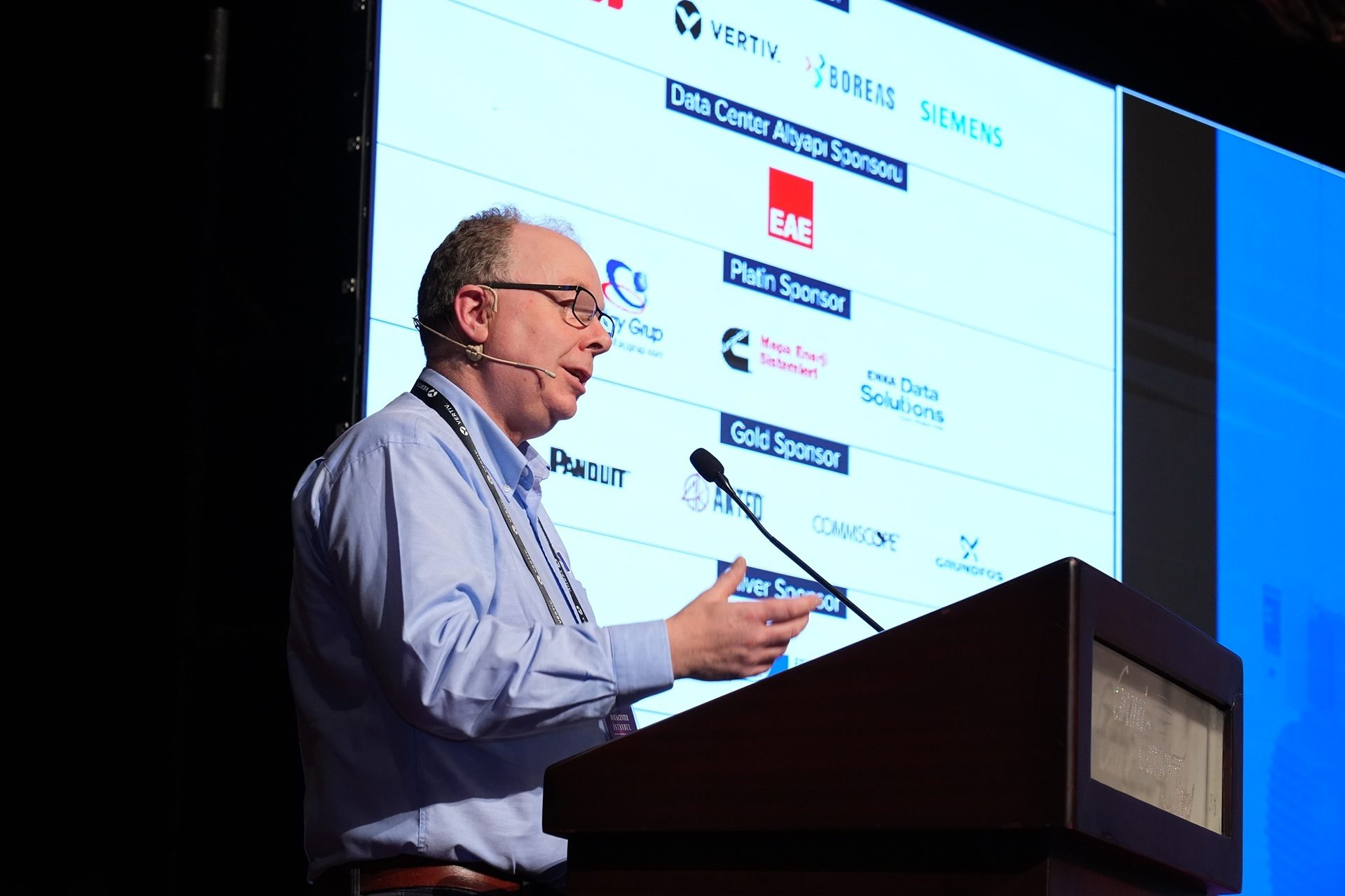 Man speaking at a podium; screen displays data center sponsors (Vertiv, Siemens, Panduit logos).