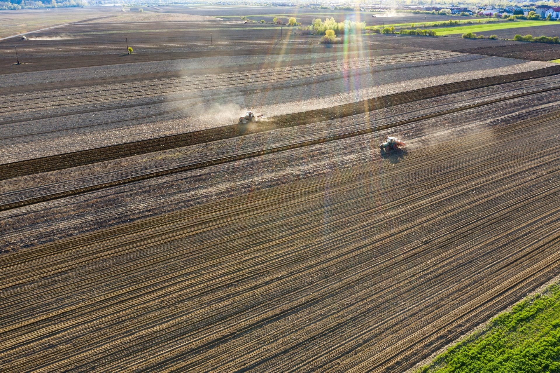 Aerial view of two tractors tilling vast agricultural fields, creating rows in the soil under bright sunlight.