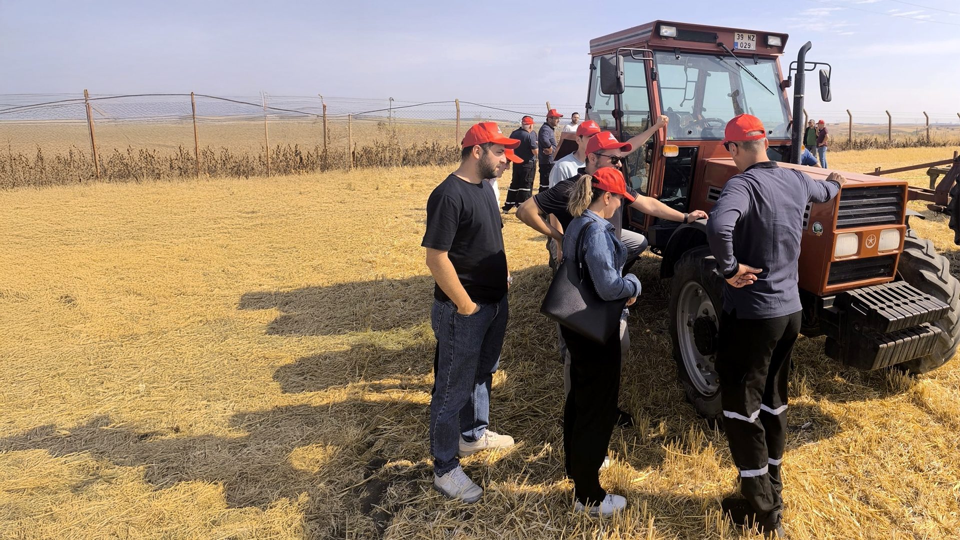 People in red caps around a brown tractor in a dry, harvested field with a fence.
