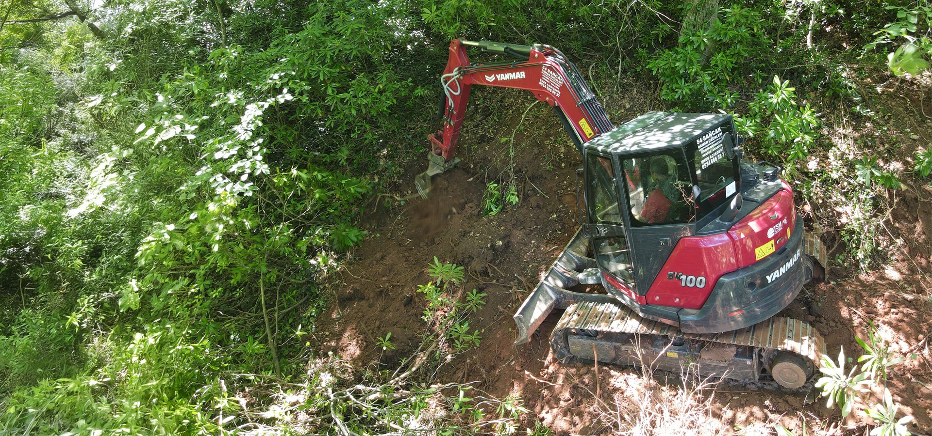 Red Yanmar SV100 excavator digging dirt on a green, vegetated slope.