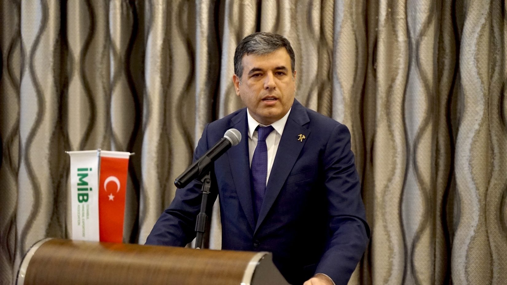 A man in a suit speaks at a podium with a microphone, flanked by IMIB and Turkish flags.