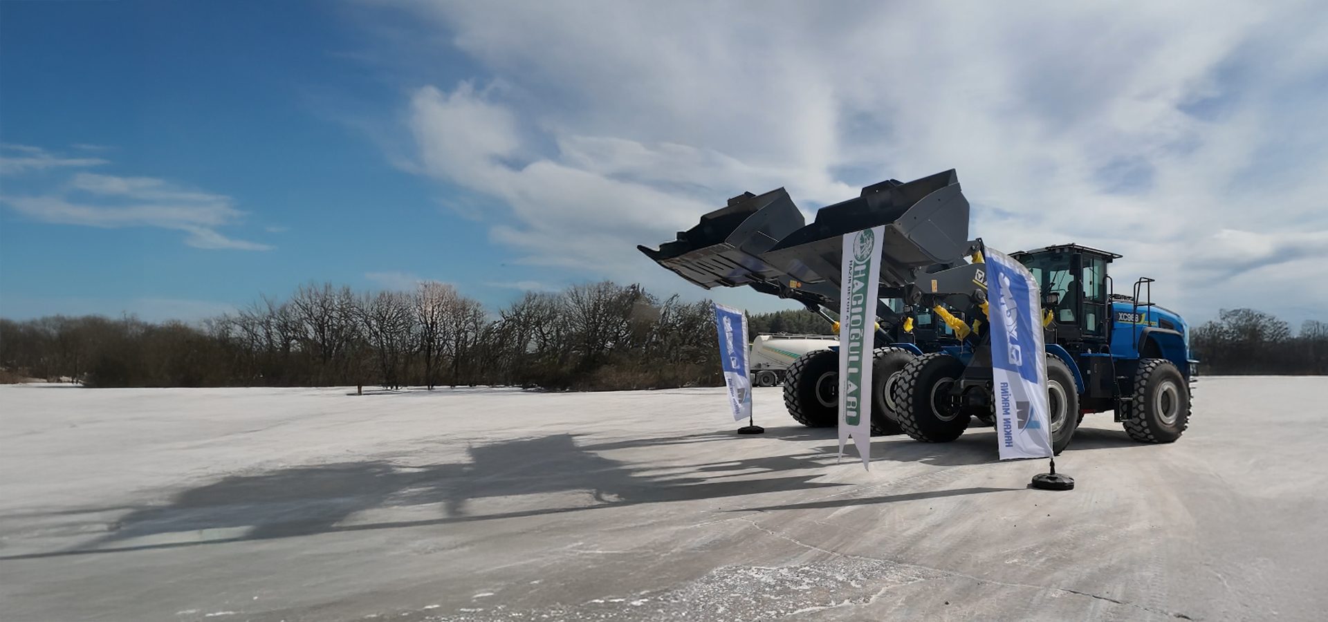 Two large blue loaders with raised buckets and flags on a frozen plain under a blue sky.
