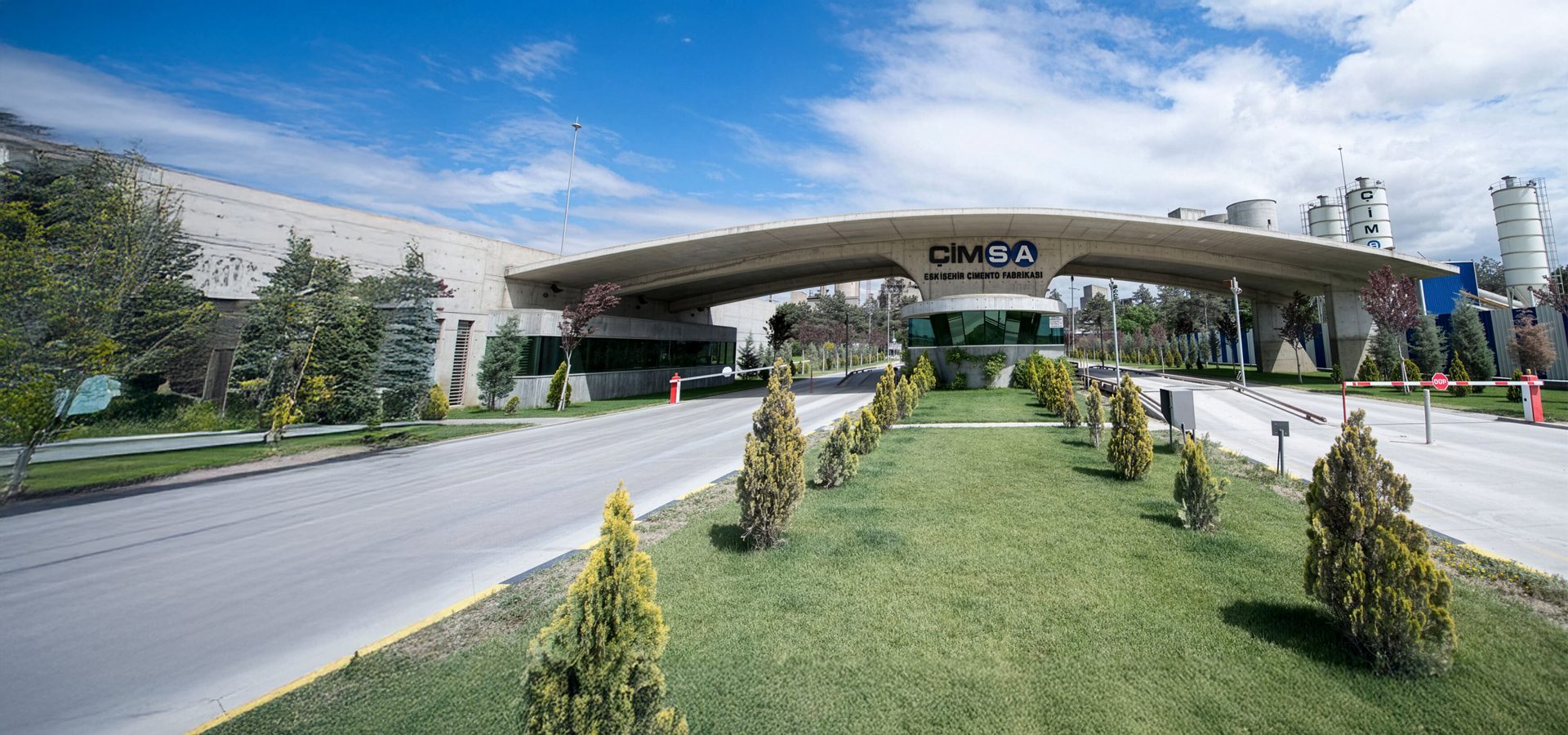 Entrance to ÇimSA Cement Factory with a large archway, security booth, and industrial silos.