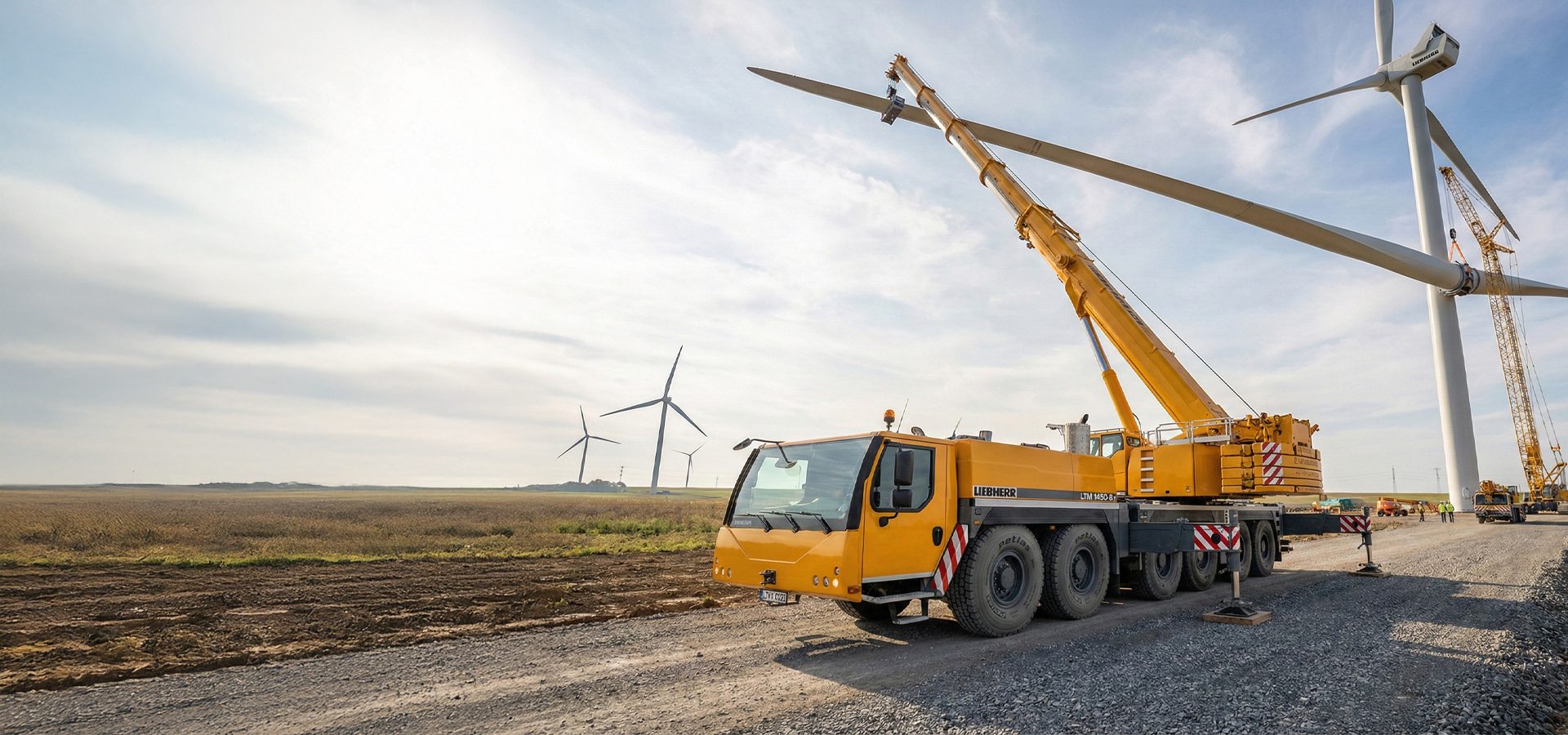 A large yellow crane lifts a wind turbine blade for installation in a field, with other turbines visible.
