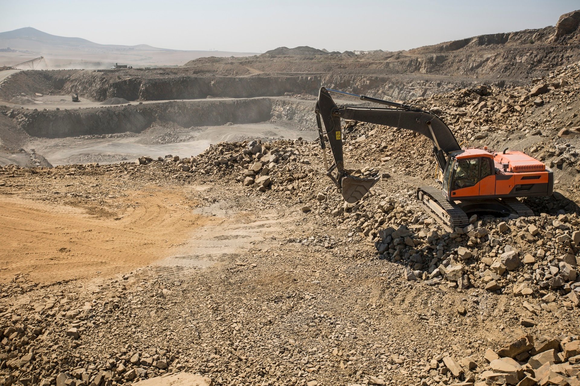 An orange and black excavator sits on a rocky, dusty surface in a multi-level open-pit quarry under a clear sky.