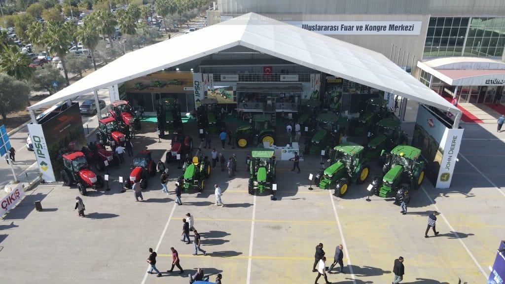 Outdoor tractor exhibition with green and red tractors under a large white tent, attended by people.