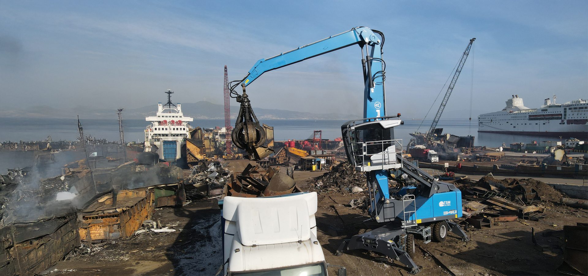 A blue material handler works at a shipbreaking yard, moving scrap metal near large vessels on a sunny day.